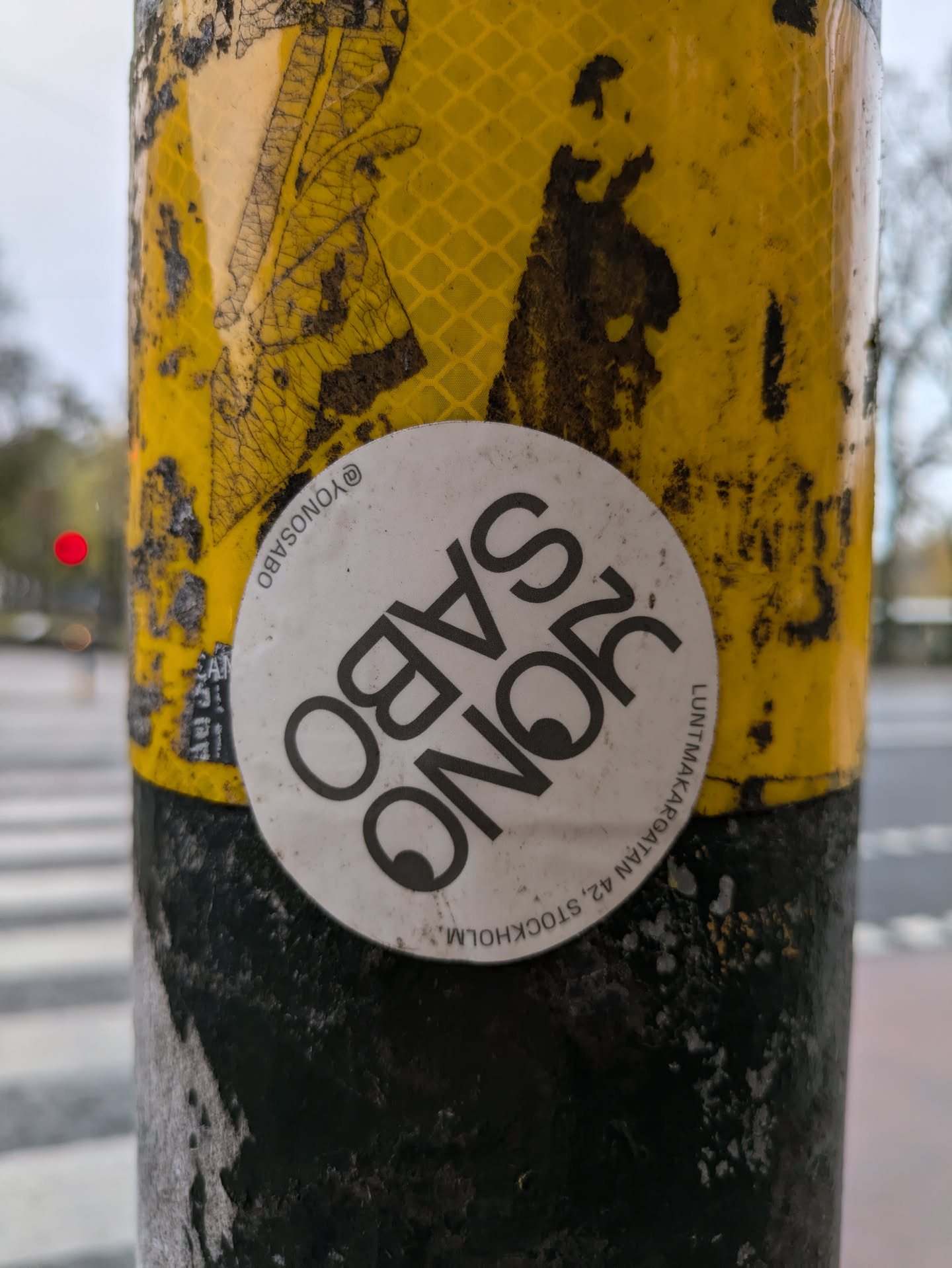 A round white sticker with black text that says 'RQNO Sabo' and an address in Stockholm. The sticker is affixed to a weathered yellow and black pole.