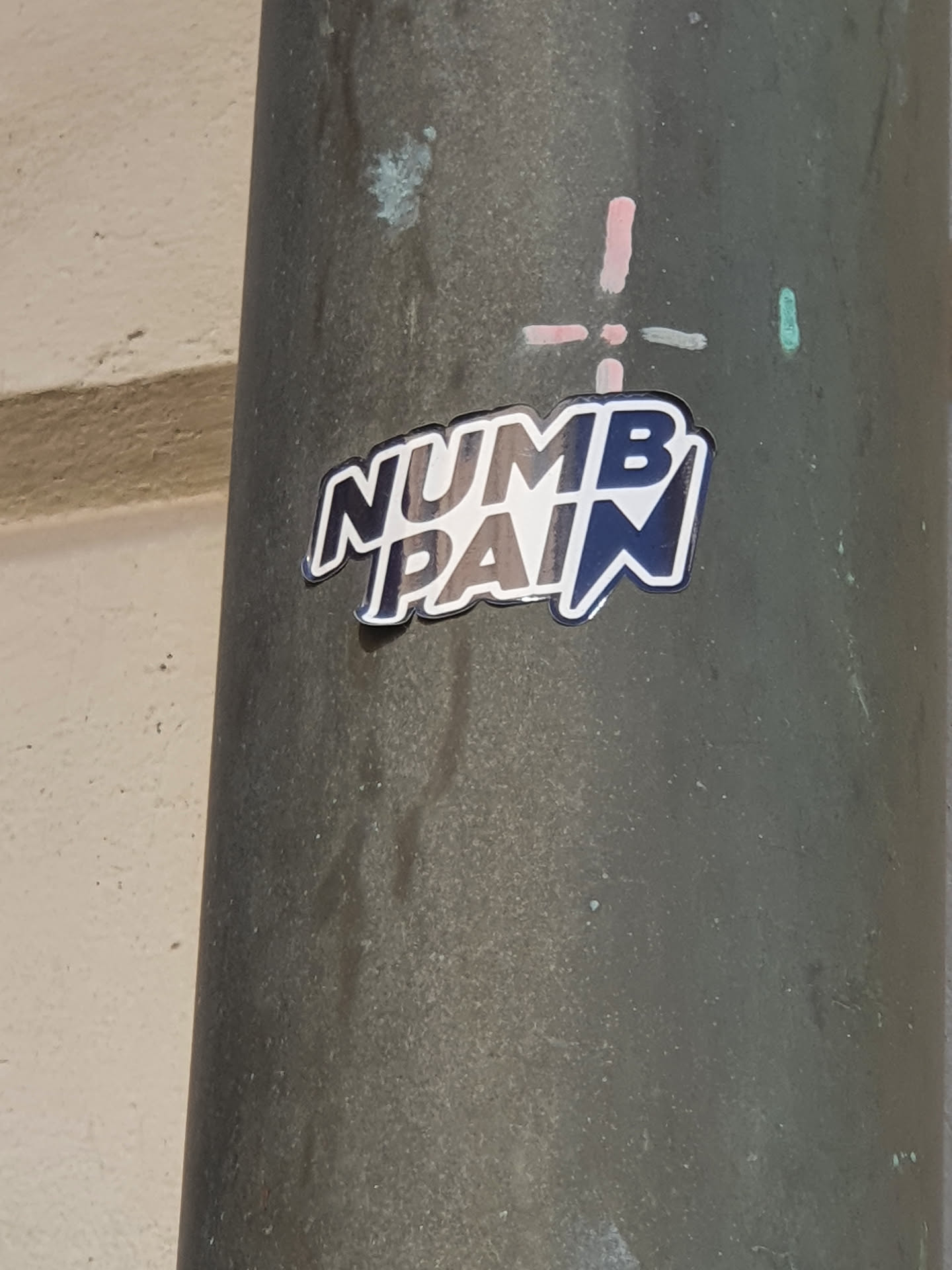 A sticker with the text "Numb Pain" in a stylized font. The sticker is dark-colored with light-colored lettering. It is affixed to a dark gray pole.