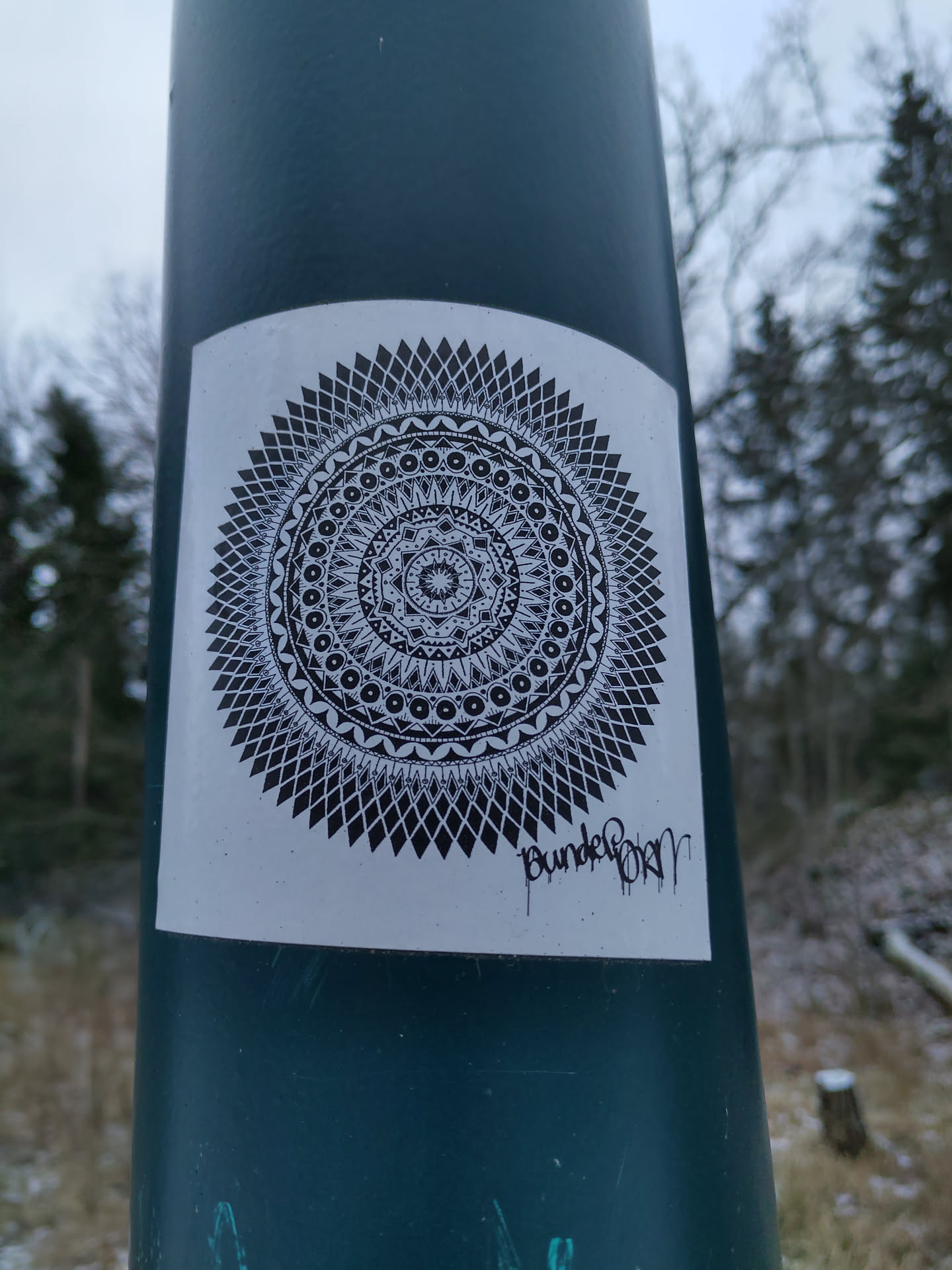 A square sticker featuring a detailed black and white mandala design. The mandala is surrounded by a geometric pattern, and the sticker is affixed to a teal pole outdoors. The artist's signature, 'Bunderborn', is visible at the bottom.