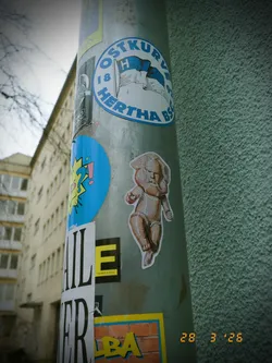 The central sticker depicts a stylized baby-like figure rendered in a mosaic art style. The figure appears to be standing or taking a step, with its arms slightly bent. Its body is composed of numerous small, irregularly shaped 'tiles' in various shades of brown, beige, and light gray, giving it a textured, almost sculptural appearance. The head is large in proportion to the body, with simplified facial features. The sticker has a white border, indicating it's a die-cut design, and is applied to a metal pole.