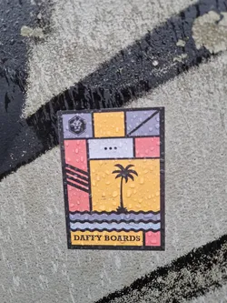 A colorful sticker featuring a geometric design with a palm tree and ocean waves. The text "DAFFY BOARDS" is visible at the bottom. The sticker appears to be wet, with water droplets visible on its surface.