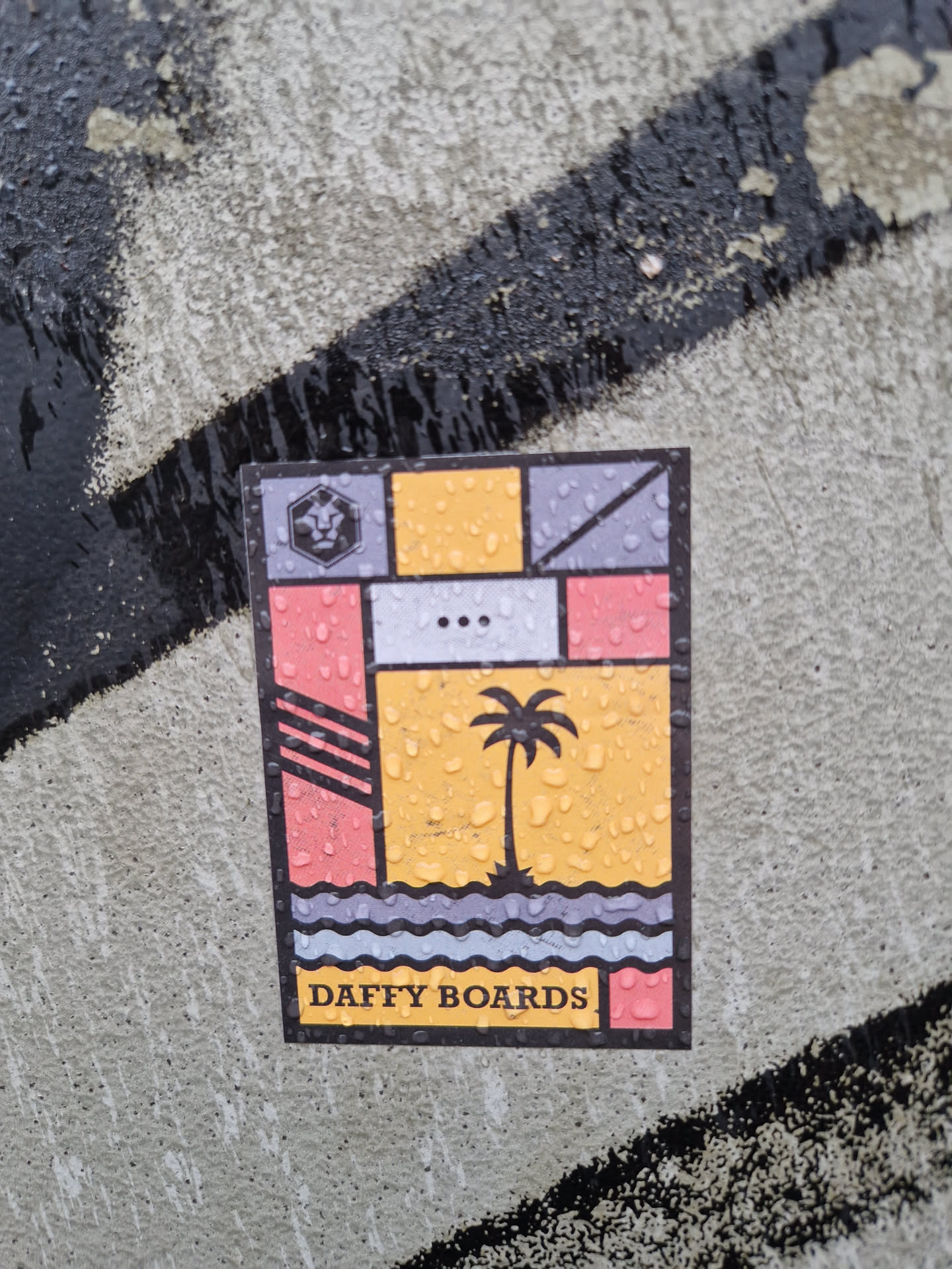 A colorful sticker featuring a geometric design with a palm tree and ocean waves. The text "DAFFY BOARDS" is visible at the bottom. The sticker appears to be wet, with water droplets visible on its surface.