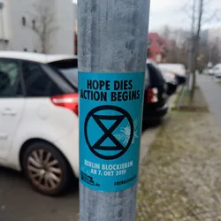 A light blue sticker with a black Extinction Rebellion symbol and text that says in English "Hope dies action begins" and in German "Berlin Blockieren ab 7. Okt 2019" which translates to "Block Berlin from October 7th, 2019". There is also a hashtag #rebelforfuture on the sticker.