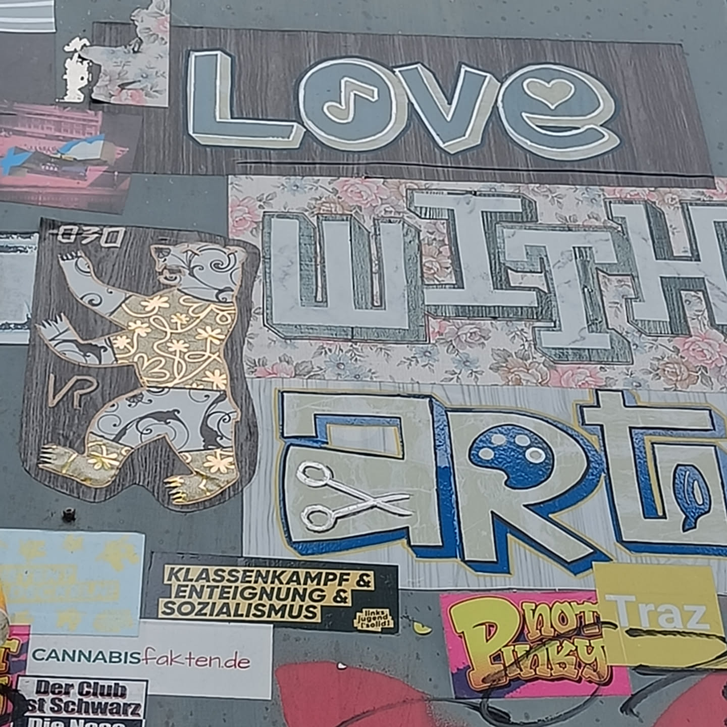 A sticker featuring a bear in a gold design. The sticker is part of a larger collection of stickers on a wall, some with political messages.  The word 'Love' with a musical note and heart, 'with' and 'Art' with scissors and a paint palette are visible on the wall around the bear sticker.