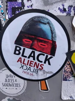 Round sticker featuring a stylized illustration of a head wearing a helmet, with the text "BLACK ALIENS" in red and black. The text is mirrored on the bottom.