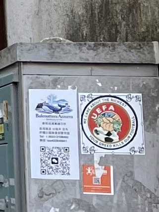 The central sticker is square with a decorative white border featuring blue floral or scroll-like patterns in each corner and along the edges. Inside this border, there is a circular emblem with a vibrant red background. This emblem parodies the UEFA logo, depicting a figure whose hands are clutching large wads of green cash, emerging from what appears to be a stylized football or globe. The letters 'UEFA' are prominently displayed in white at the top of the red circle. Arcing around the red circle, within the white border, is text that reads 'WE CARE ABOUT THE RICHEST' at the top and 'YOUR GREED KILLS OUR FOOTBALL' at the bottom. The overall color palette includes red, white, blue, and green.