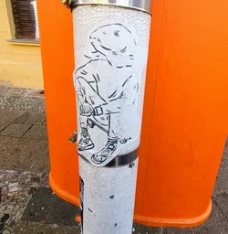 This sticker depicts a stylized character with the head of a fish and the body of a person. The character is depicted sitting on what appears to be a skateboard. The sticker is applied to a weathered metal pole and partially obscures another sticker underneath.