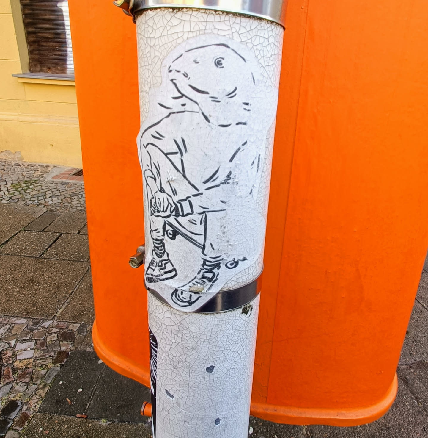 This sticker depicts a stylized character with the head of a fish and the body of a person. The character is depicted sitting on what appears to be a skateboard. The sticker is applied to a weathered metal pole and partially obscures another sticker underneath.
