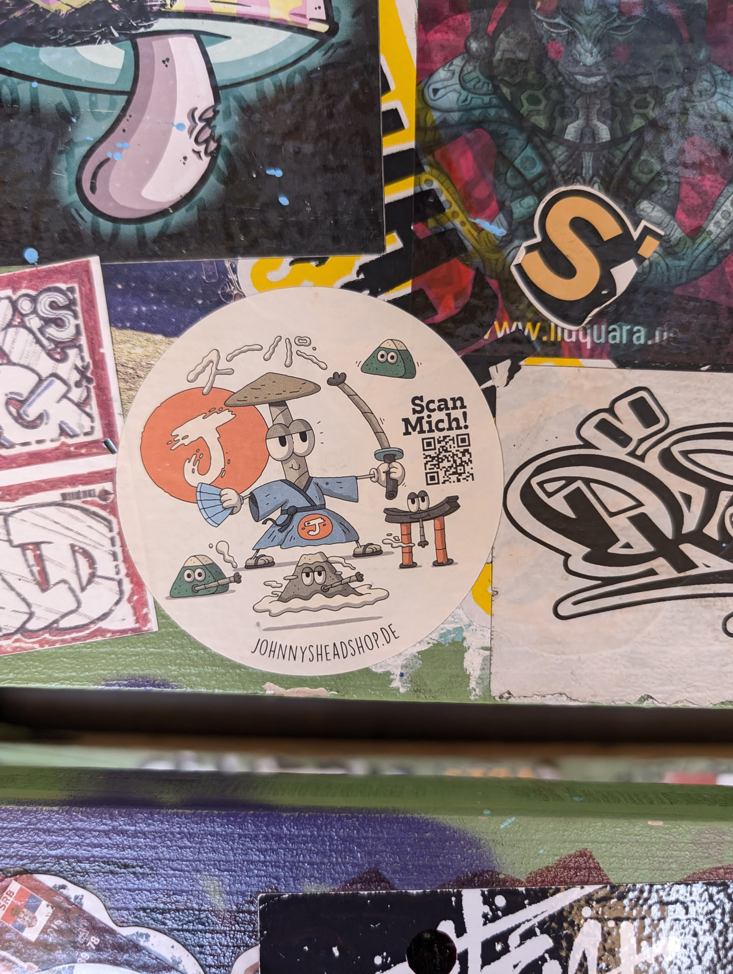 Round sticker featuring an illustration of a cartoon character in a samurai outfit, holding a sword. Other elements include onigiri, rocks, and a QR code with the text "Scan Mich!"  The sticker also has Japanese text and the website "johnnysheadshop.de".