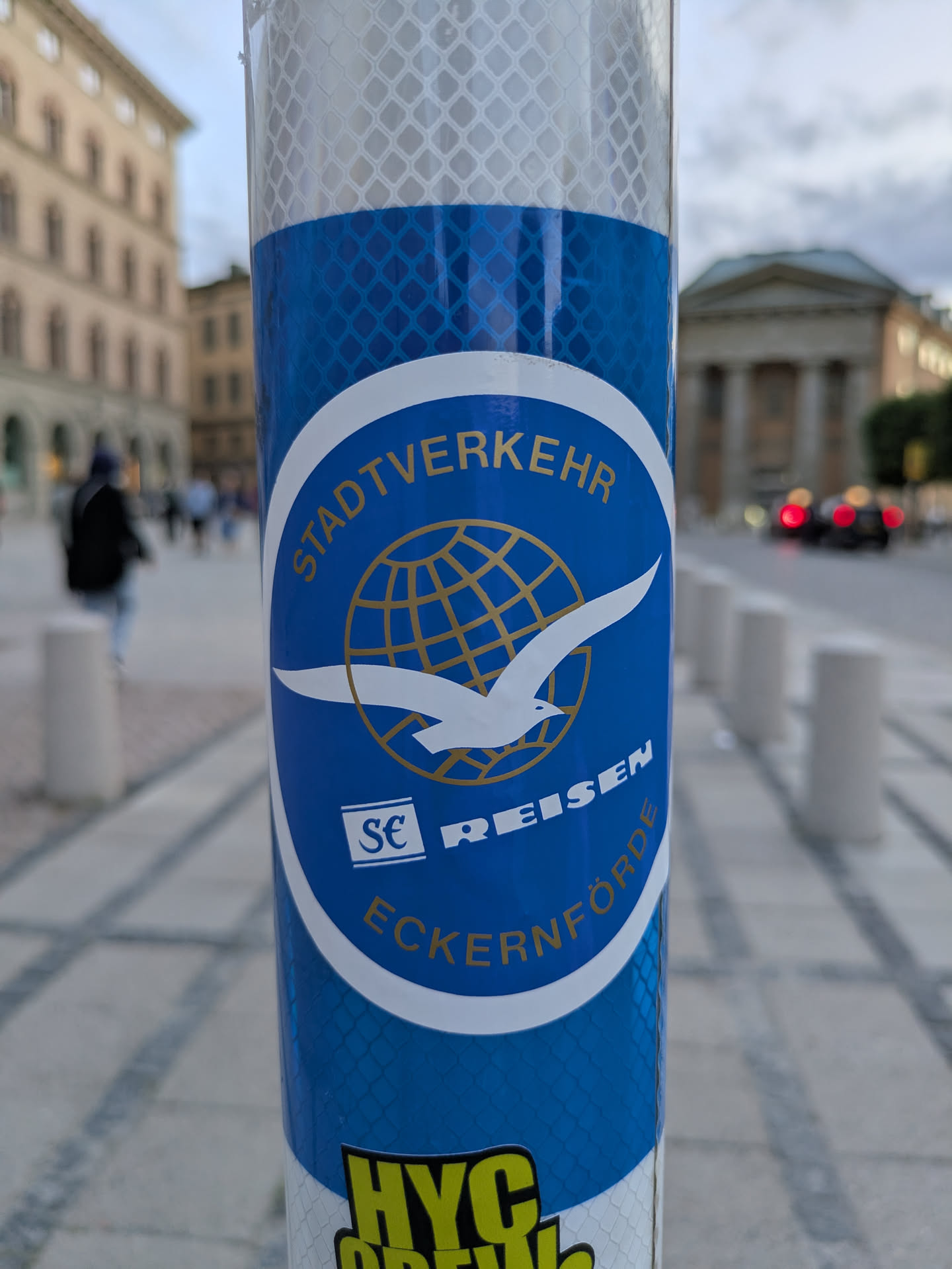 The sticker is predominantly blue and white with gold lettering. It features a circular emblem at its center. Within the circle, a stylized white seagull is depicted in flight, with its wings spread. Behind the seagull is a golden globe with a grid pattern representing longitude and latitude lines. The upper arc of the circle contains the text 'STADTVERKEHR' in gold capital letters. Below the seagull and globe, the words 'REISEN' and 'ECKERÜTFÖRDE' are also present in gold, with 'REISEN' on a white background and 'ECKERÜTFÖRDE' on a blue background. The entire sticker is affixed to a reflective, diamond-patterned surface, likely a pole.