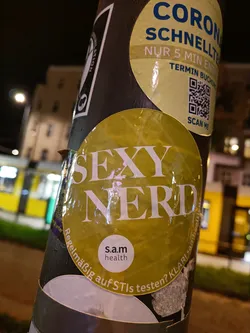 A yellow sticker with the text "SEXY NERD" in bold white letters. There is a smaller, circular sticker in the bottom featuring s.a.m health logo and text. The main sticker is affixed to a pole with other stickers.  One of them is a QR code for Corona Schnelltest. A tram is visible in the background.