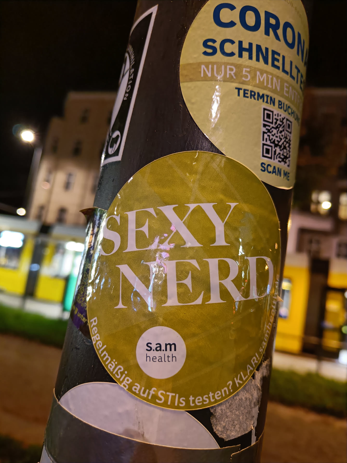 A yellow sticker with the text "SEXY NERD" in bold white letters. There is a smaller, circular sticker in the bottom featuring s.a.m health logo and text. The main sticker is affixed to a pole with other stickers.  One of them is a QR code for Corona Schnelltest. A tram is visible in the background.