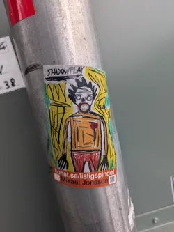 A colorful sticker of an image that looks like the style of Basquiat. The sticker is on a metal pole and has the text "SHADOWPLAY" and "konst.se/listigspindel Mikael Jonsson" on it.