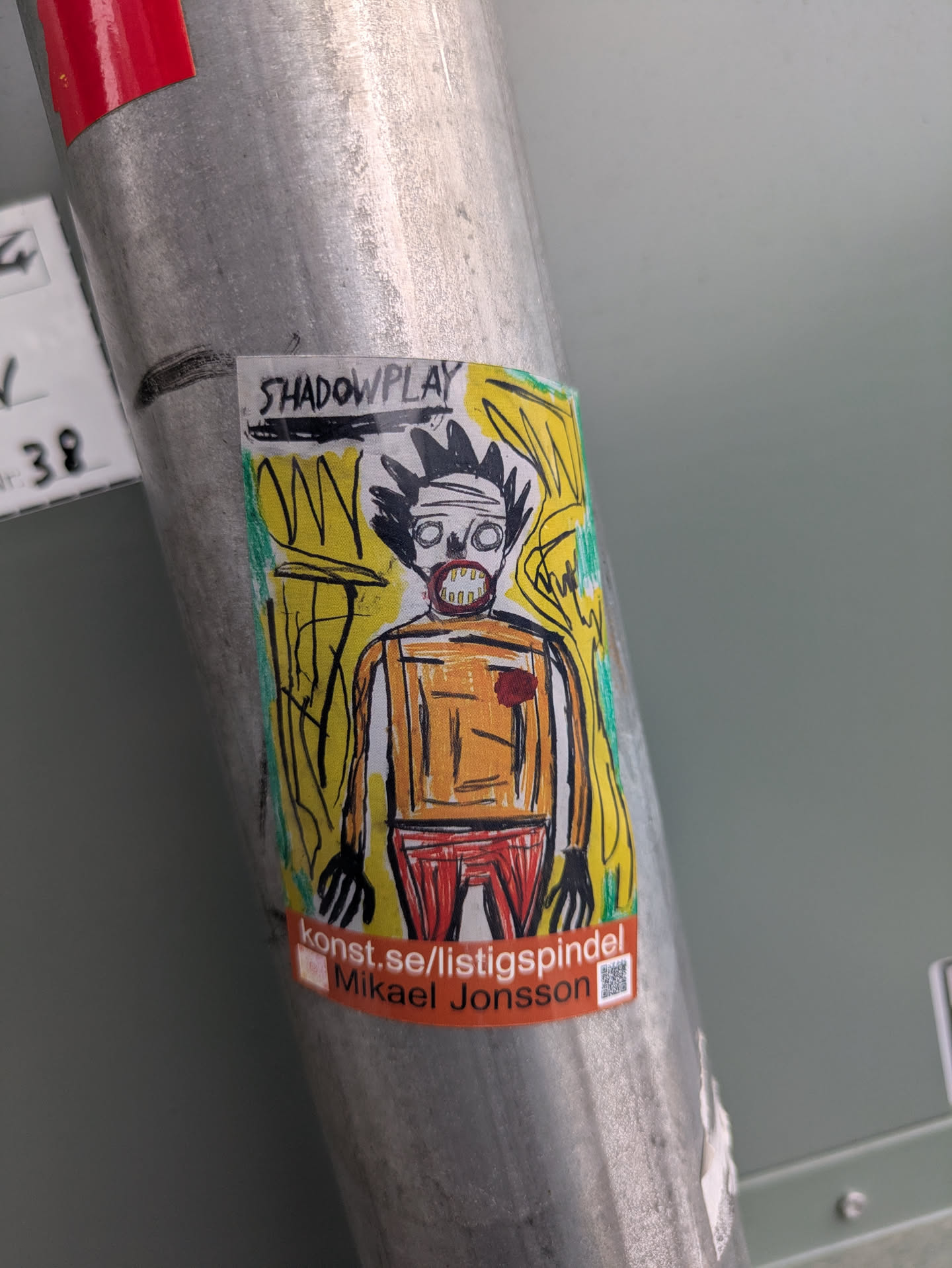 A colorful sticker of an image that looks like the style of Basquiat. The sticker is on a metal pole and has the text "SHADOWPLAY" and "konst.se/listigspindel Mikael Jonsson" on it.