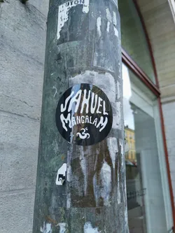 Black and white round sticker with the text JAHUEL Mangalam and an Om symbol. The sticker is affixed to a weathered pole.