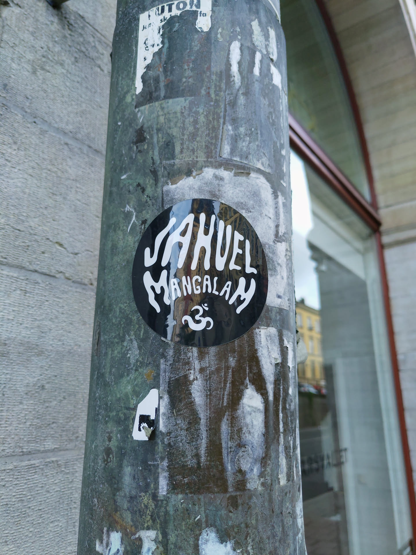 Black and white round sticker with the text JAHUEL Mangalam and an Om symbol. The sticker is affixed to a weathered pole.