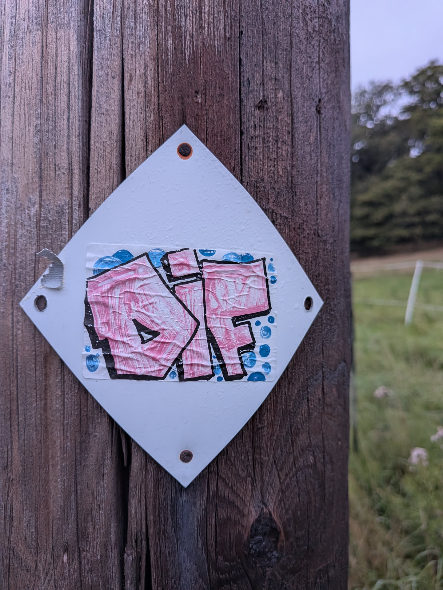 The sticker is a roughly square piece of paper with a white, diamond-shaped background, affixed to a wooden post with two small rivets. The sticker itself features prominent graffiti lettering in a bold, bubble-style font. The letters, appearing to spell 'DIF' or a similar variation, are filled with a textured pink color and outlined in thick black. Scattered around the lettering are small, irregular blue shapes that resemble bubbles or water droplets. The sticker shows signs of wear, with some creasing and slight peeling at the edges, particularly on the left side where a piece of tape is also applied.