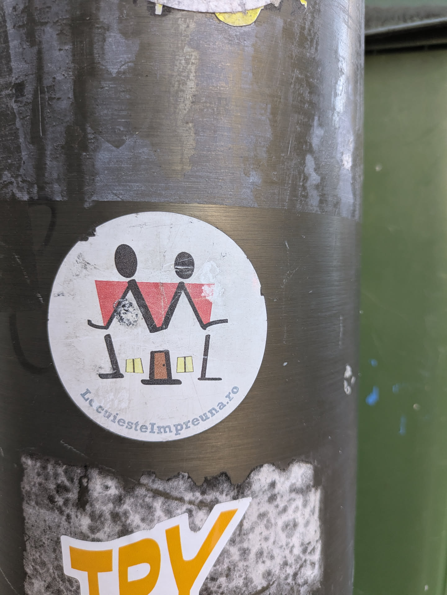 A round sticker on a pole features an illustration of a house with two stick figures holding it up. Below the image, the text "LocuiesteImpreuna.ro" is printed. The style is minimalist and playful, suggesting a message related to living together or community.