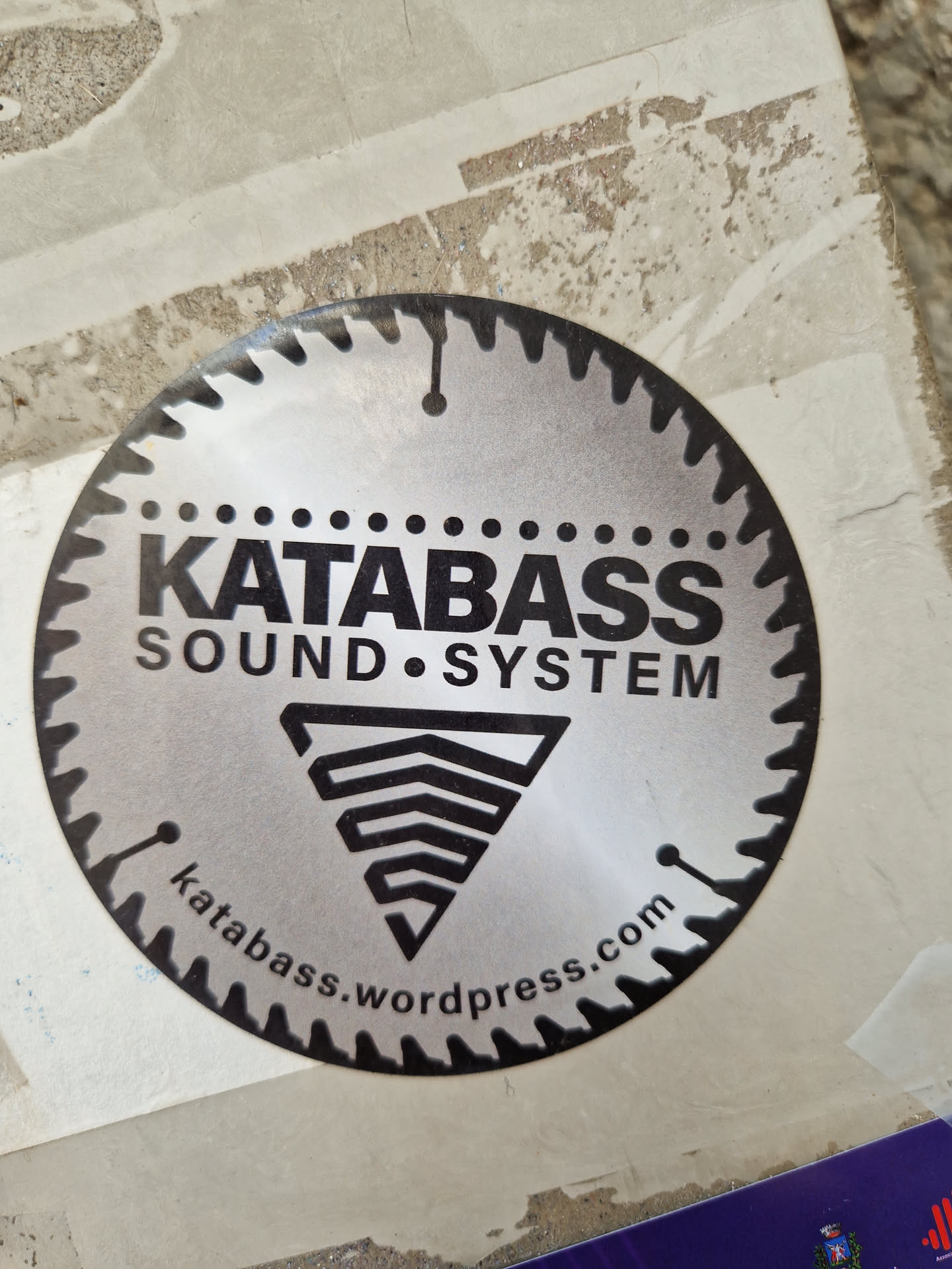 A round sticker with a saw blade design. It features the text "KATABASS SOUND SYSTEM" and a logo. The website katabass.wordpress.com is also printed on the sticker.
