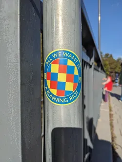 A round sticker with a checkerboard pattern in the center and text surrounding it. The text says "All we want is a running riot". The years 86 and 91 are written on the sides of the circle. The sticker is attached to a metal pole.