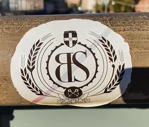 A round sticker featuring a crest-like design with the letters "BS" prominently displayed within a circular frame. The crest is flanked by wheat stalks, and includes a smaller emblem at the bottom with crossed keys. The overall aesthetic is vintage and reminiscent of a brewery logo.