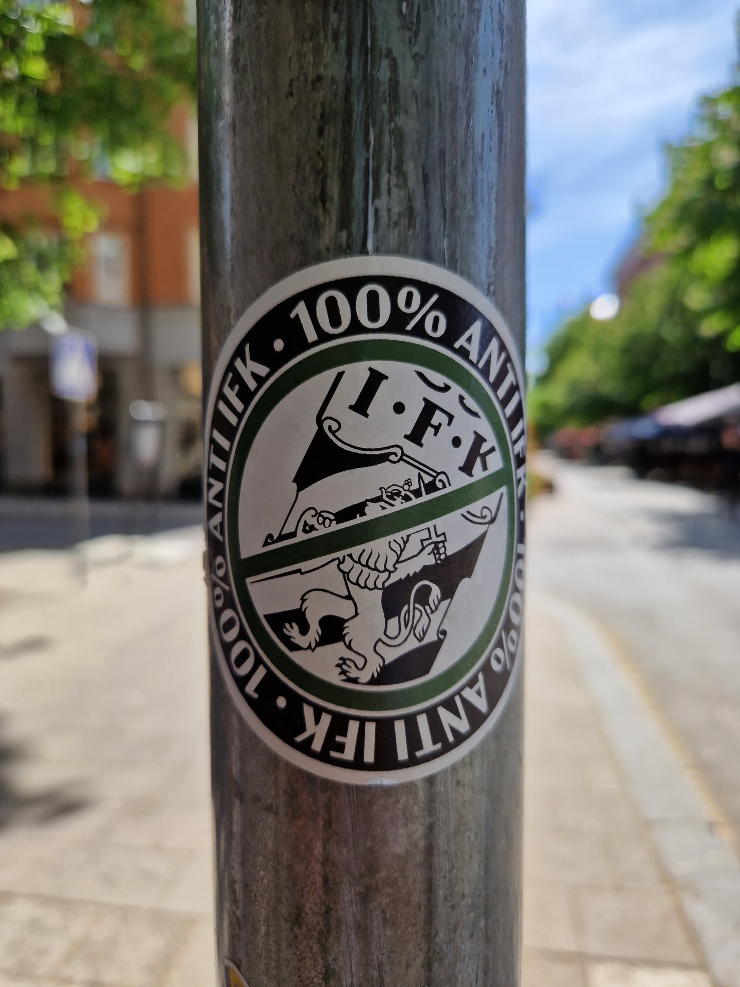 A round sticker with the text "100% Anti IFK" written in a circle.  The inner circle features a graphic of a lion emblem crossed out. The sticker is affixed to a metal pole.
