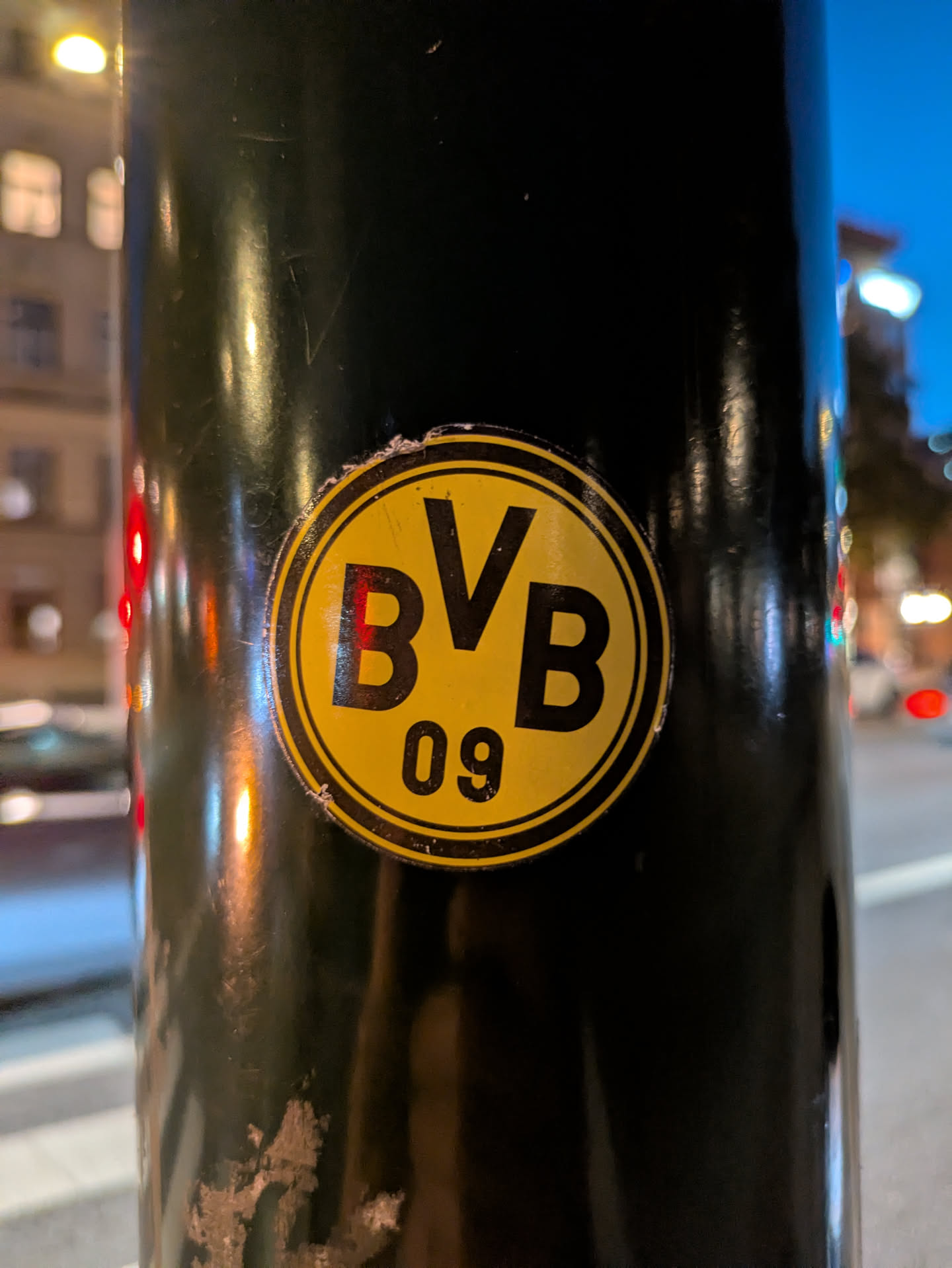 A circular sticker prominently affixed to a dark, possibly black or dark green, metal pole. The sticker features a bright yellow background with bold black text. The letters 'BVB' are centrally placed in a large font, with a smaller '09' positioned directly below them. A thin black ring outlines the inner yellow circle, and a thicker black ring forms the outer border of the circular sticker. The sticker shows minor wear around its edges, and reflections from surrounding lights are visible on its glossy surface and the pole.