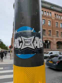Street sticker A rectangular sticker placed horizontally on a dark metal street pole, partially covering a circular blue sticker beneath it. The sticker features the bold acronym 'ACAB' styled in heavy block letters, with a central four-pointed starburst badge separating the letters 'AC' and 'AB'. Inside the badge is a small crest inscribed with the letters 'DIF', representing the Swedish sports club Djurgårdens IF. The sticker is rendered in black, white, and subtle grey tones with weathered texture effects.