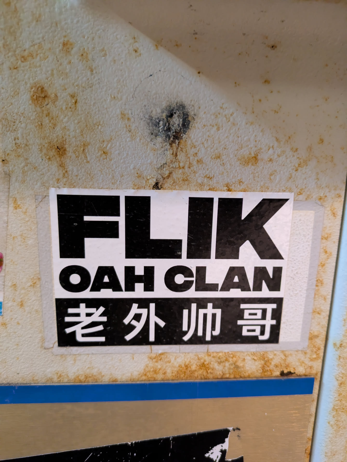 This is a black and white sticker with the text "FLIK OAH CLAN" in bold letters on a white background. Under that there is a text in Chinese "老外帅哥". The wall is discolored and shows signs of wear.