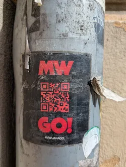 A black square sticker on a pole featuring the text 'MW' stacked on top of a QR code. Below the QR code is the word 'GO!' in red, followed by the hashtag '#MWUANAGO'.