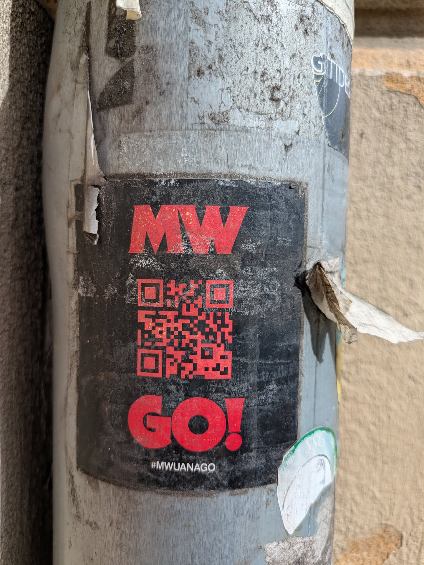 A black square sticker on a pole featuring the text 'MW' stacked on top of a QR code. Below the QR code is the word 'GO!' in red, followed by the hashtag '#MWUANAGO'.