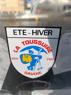 Vintage sticker from La Toussuire, a ski resort in the Savoie region of France.  The sticker shows a cartoon skier with a sun on its chest. It indicates that the resort is open in both summer and winter, and specifies an altitude range of 1800 to 2400 meters.