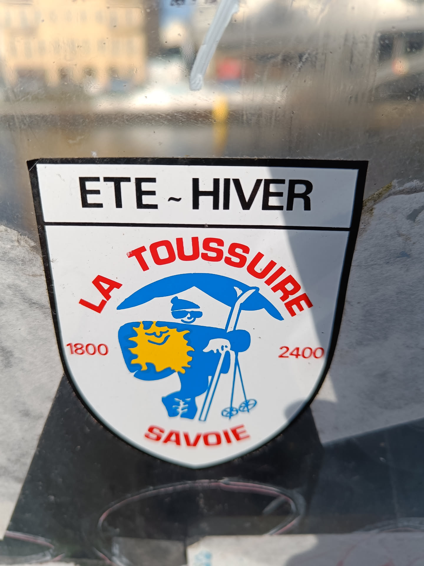 Vintage sticker from La Toussuire, a ski resort in the Savoie region of France.  The sticker shows a cartoon skier with a sun on its chest. It indicates that the resort is open in both summer and winter, and specifies an altitude range of 1800 to 2400 meters.