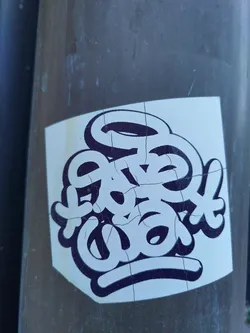 A sticker featuring stylized lettering in black and white. The lettering appears to be hand-drawn or graffiti-inspired. The sticker is slightly worn and shows some cracks, giving it a lived-in feel. It's been affixed to a gray pole outdoors.