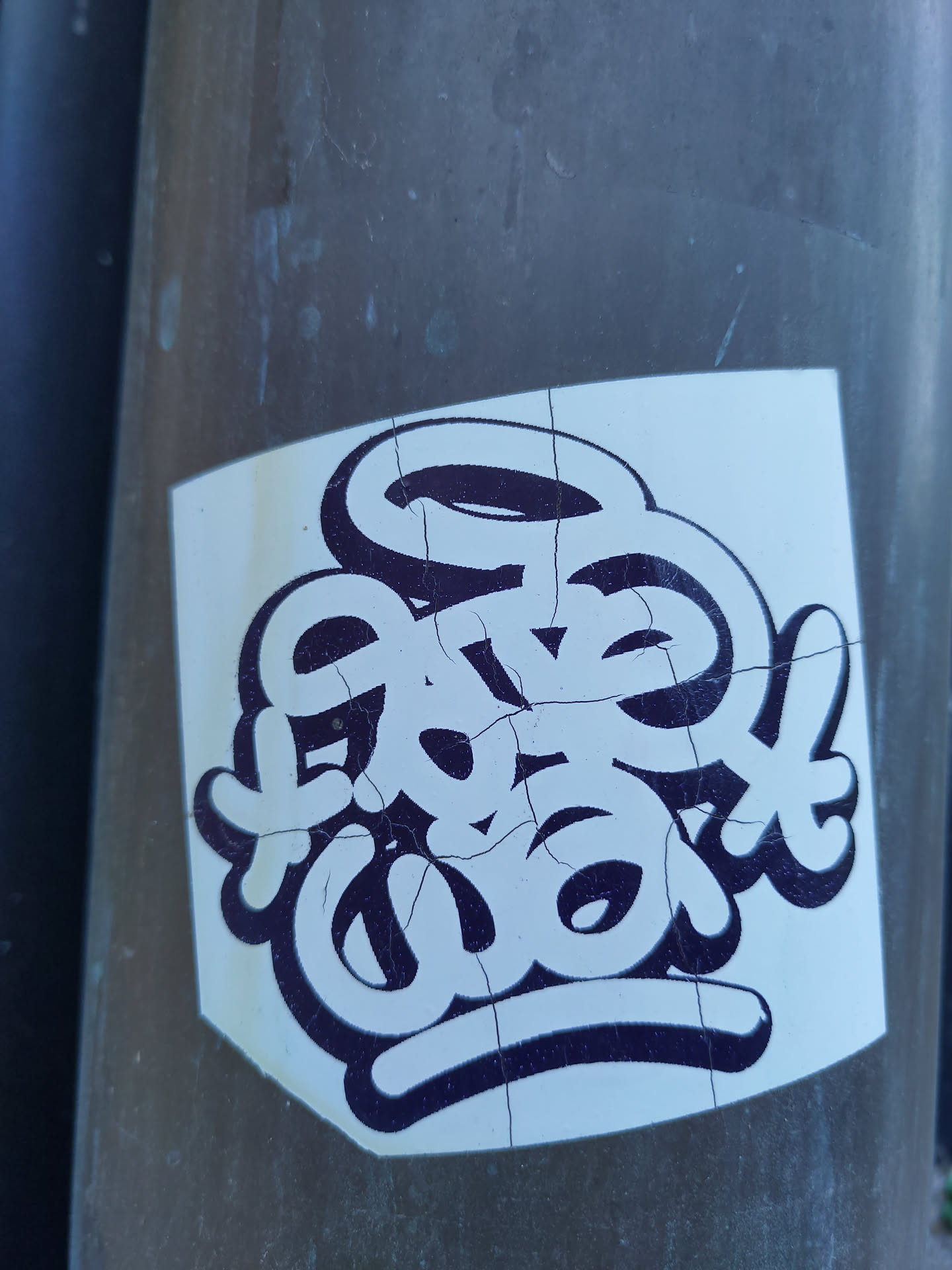 A sticker featuring stylized lettering in black and white. The lettering appears to be hand-drawn or graffiti-inspired. The sticker is slightly worn and shows some cracks, giving it a lived-in feel. It's been affixed to a gray pole outdoors.