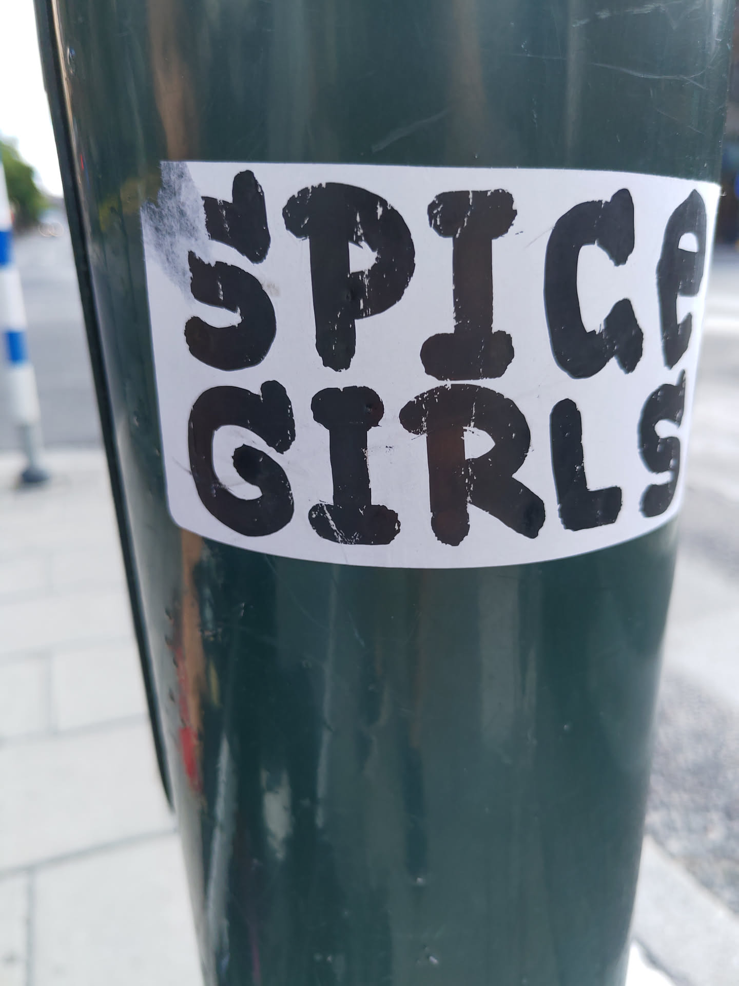 A sticker featuring the words "Spice Girls" in a bold, slightly distressed black font on a white background.  The sticker is affixed to a dark green pole.