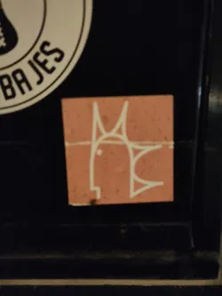 A small, peach-colored sticker with a minimalist line drawing of a fish. The fish is white and has a simple, almost geometric design. The sticker is affixed to a dark surface.