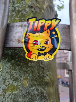 A yellow cat with pink eyes wearing a bow tie. The sticker is slightly crumpled and attached to a tree trunk.  The name "TIPPY" is written above the cat in stylized lettering.