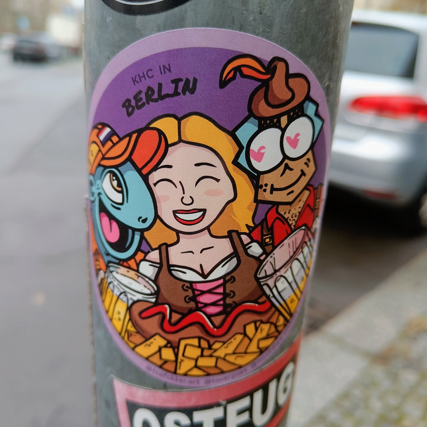 A round sticker featuring a cartoon illustration of three characters enjoying beer and fries.  The characters include a woman in traditional German attire, a blue creature, and a man in a hat. The background is purple, and the text 'KHC IN BERLIN' is visible at the top.