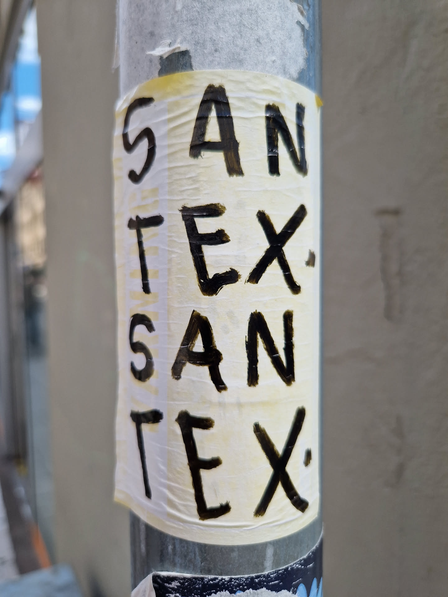 A sticker with the words 'SAN TEX.' written in black marker on a off-white background. The sticker is slightly wrinkled and is affixed to a grey metal pole.