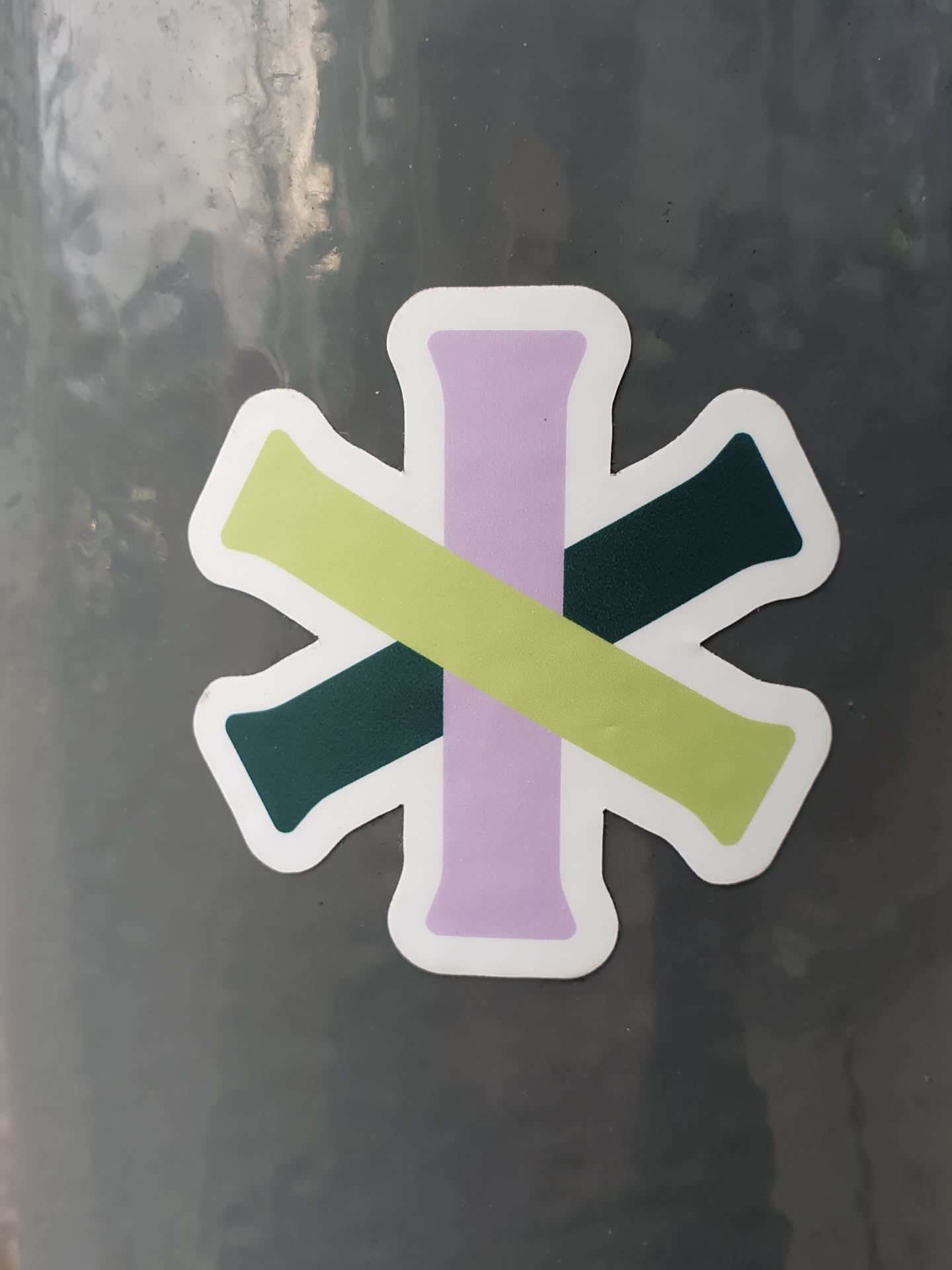 Close-up view of a sticker featuring an abstract design. The sticker consists of four rectangular shapes in pale purple, light green, and dark green, arranged to form a cross-like pattern. The shapes overlap and intersect, creating a visually interesting composition. The sticker has a white border.