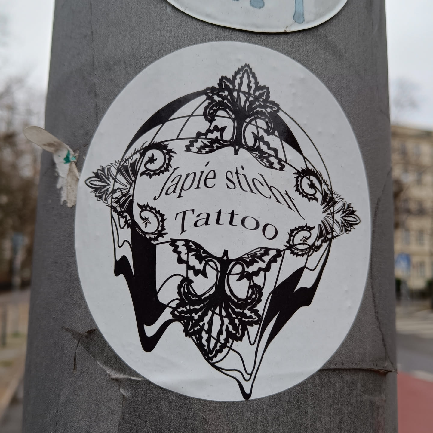 Black and white sticker with intricate design featuring a globe, butterflies, and floral elements.  The text "Japie Stich Tattoo" is prominently displayed.