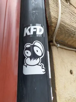 A black and white sticker featuring a simple cartoon character and the letters KFD.  The sticker is applied to a dark grey drainpipe.