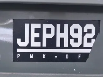 A black and white sticker with the text 'JEPH92' in a bold, uppercase sans-serif font. Below the main text, there are smaller letters: 'P M K - D F'. The sticker is slightly torn at the bottom right corner and is adhered to a dark gray surface.