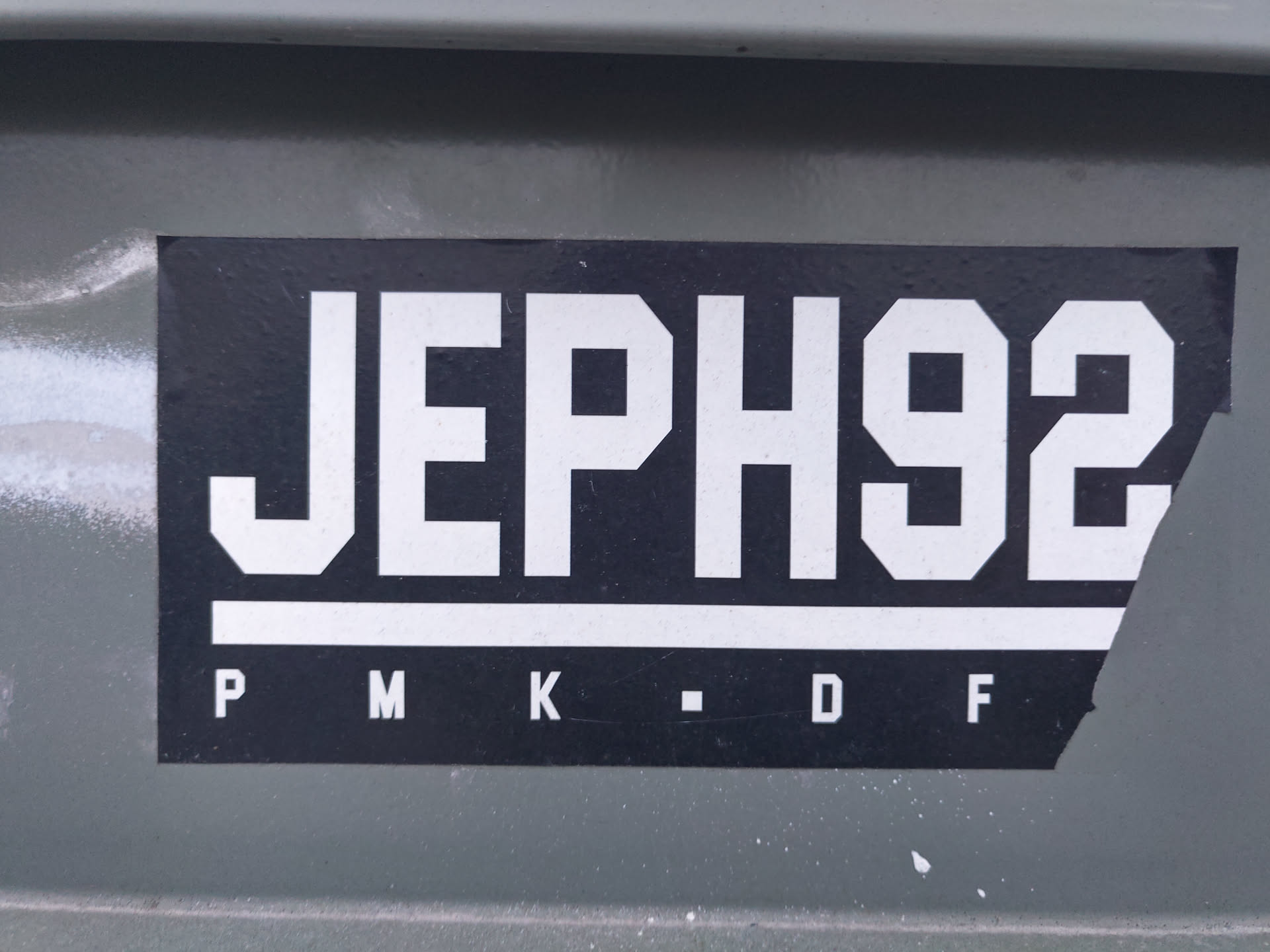 A black and white sticker with the text 'JEPH92' in a bold, uppercase sans-serif font. Below the main text, there are smaller letters: 'P M K - D F'. The sticker is slightly torn at the bottom right corner and is adhered to a dark gray surface.