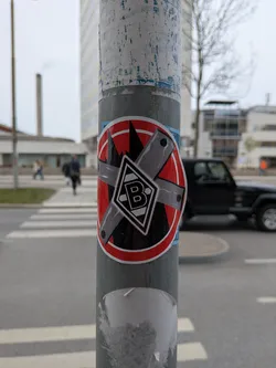 A circular sticker with a prominent red border, featuring a serrated inner edge. The central design is set against a black background with a jagged, mountain-like top edge. Two textured grey bars, resembling metal or tape, are crossed over each other, forming an 'X' shape, indicative of a 'forbidden' or 'no entry' symbol. Each bar has small black circular details at its ends. At the center of the 'X' is a white diamond shape with a black outline, containing a stylized black capital letter 'B'. This central emblem is the logo of the German football club Borussia Mönchengladbach.