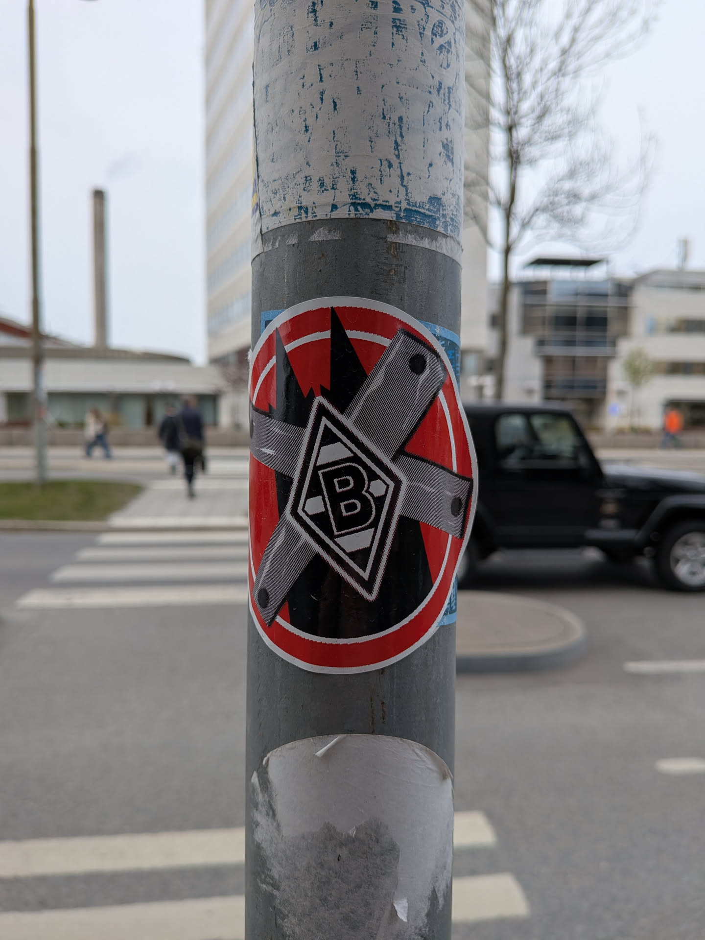 A circular sticker with a prominent red border, featuring a serrated inner edge. The central design is set against a black background with a jagged, mountain-like top edge. Two textured grey bars, resembling metal or tape, are crossed over each other, forming an 'X' shape, indicative of a 'forbidden' or 'no entry' symbol. Each bar has small black circular details at its ends. At the center of the 'X' is a white diamond shape with a black outline, containing a stylized black capital letter 'B'. This central emblem is the logo of the German football club Borussia Mönchengladbach.
