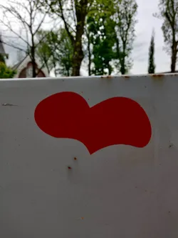 A single, hand-drawn style red heart sticker is affixed to a light gray, slightly textured surface, possibly metal. The background is blurred, showing trees and a building in the distance.
