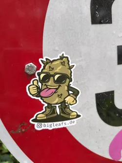 A sticker featuring a cartoon cannabis bud character wearing sunglasses and giving a thumbs up. The character has a playful expression and is wearing sneakers. The sticker has a website address '@bigleafs.de' printed on the bottom.