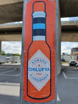A sticker featuring a hot sauce bottle design with the text "Edward Chilufya", "Tiger", and "Djurgården". The sticker is affixed to a metal pole.